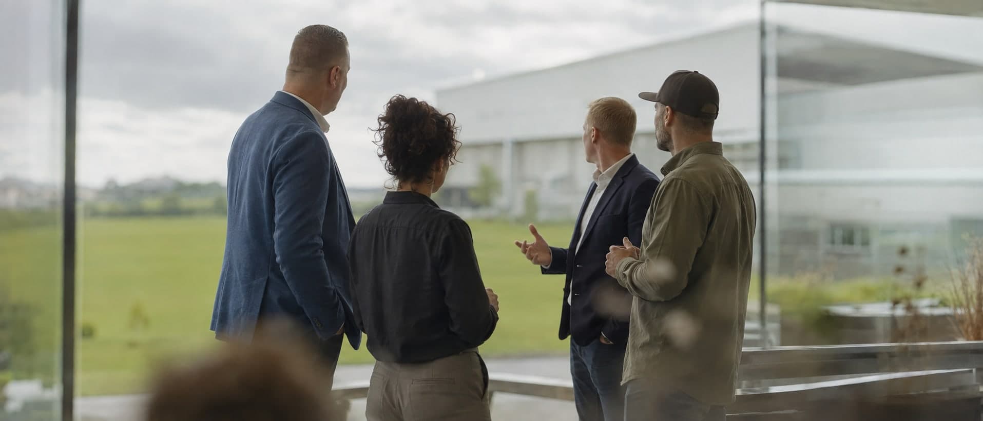 Four people in conversation at a window, looking out over a field towards a warehouse